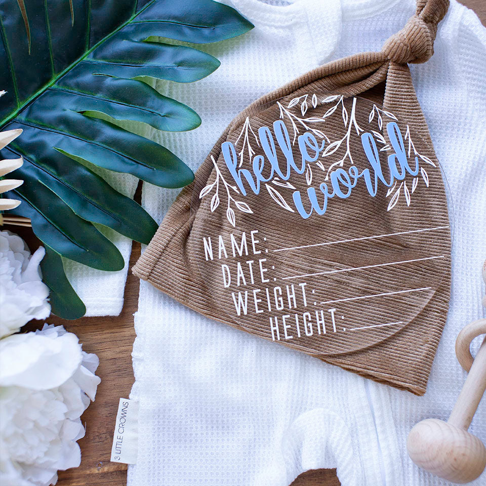 Top view of Hello Fern clear acrylic 'Hello Word' birth announcement plaque with blue wording and spaces for baby's name, birth date, weight, and height lying on baby boy onesie and beanie.