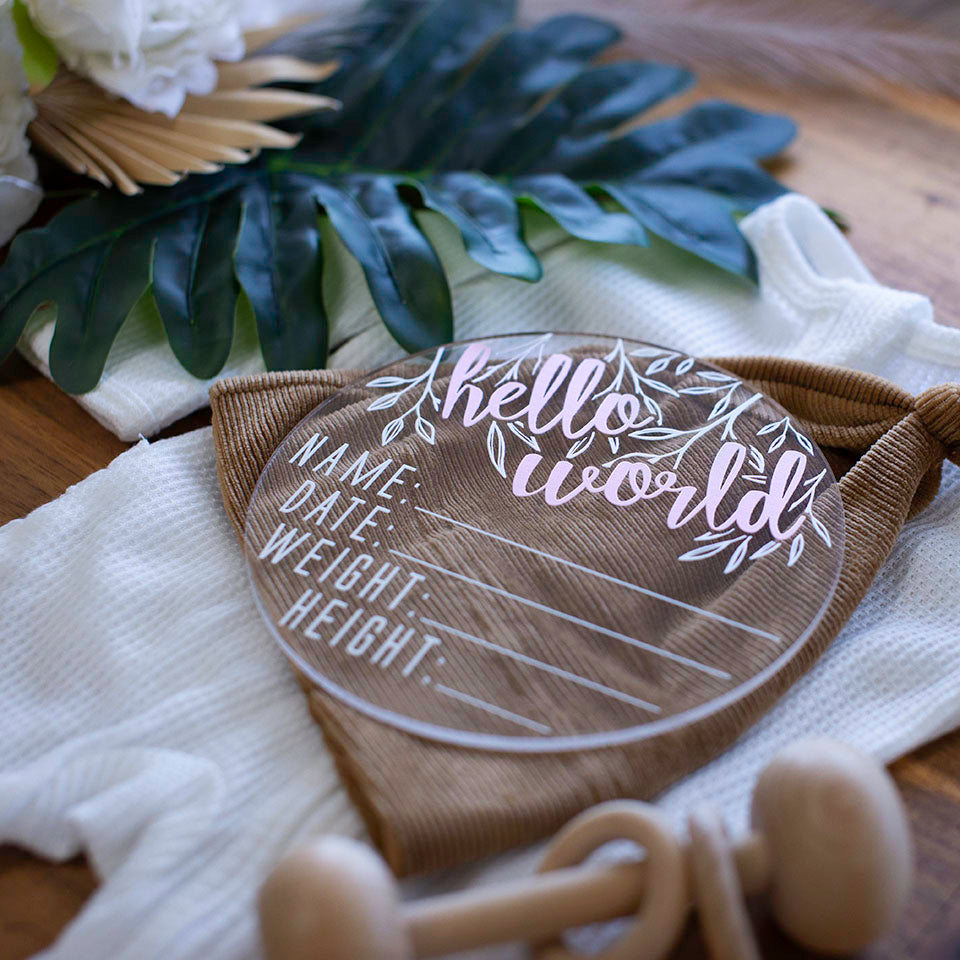 Closeup view of Hello Fern clear acrylic 'Hello Word' birth announcement plaque with pink wording and blank spaces for baby's name, birth date, weight, and height lying on baby items.