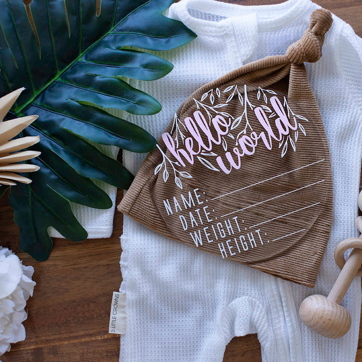 Top view of Hello Fern clear acrylic 'Hello Word' birth announcement plaque with pink wording and blank spaces for baby's name, birth date, weight, and height lying on white baby onesie and brown beanie.