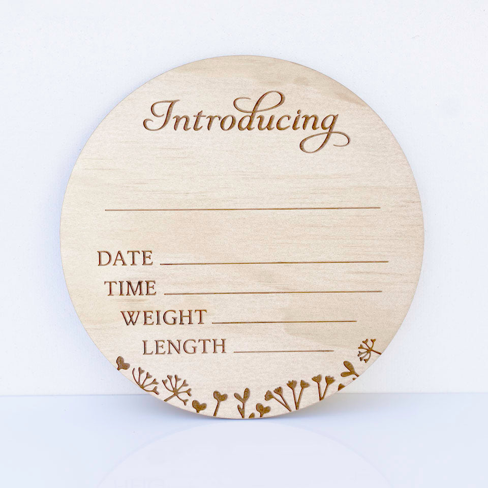 A round light wood baby birth announcement plaque with engraved text reading 'Introducing' and fields for date, time, weight, and length, plus small floral line illustrations along the bottom edge, shown on a clean white background.