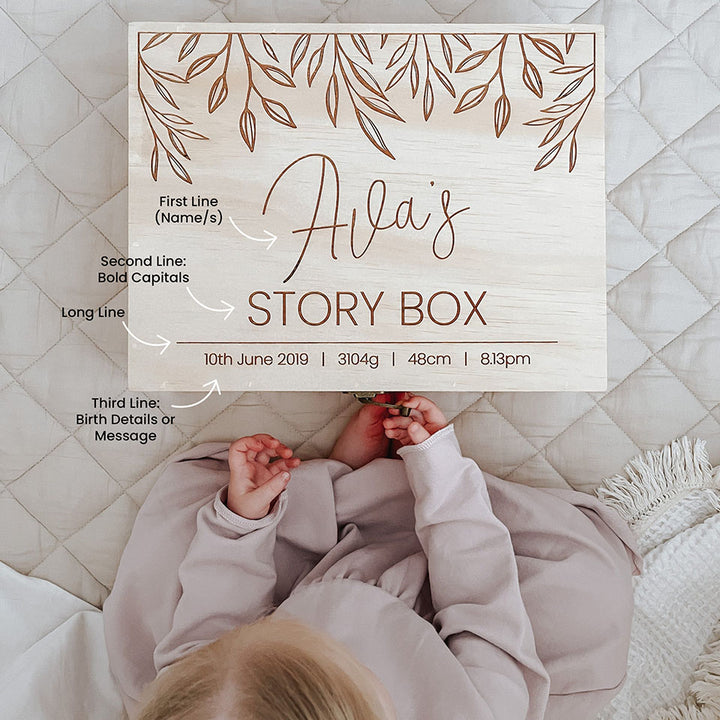 A Hello Fern personalised large wooden keepsake box labeled 'Ava's STORY BOX' with labels, shown on a quilted cream bed with a baby lying underneath.