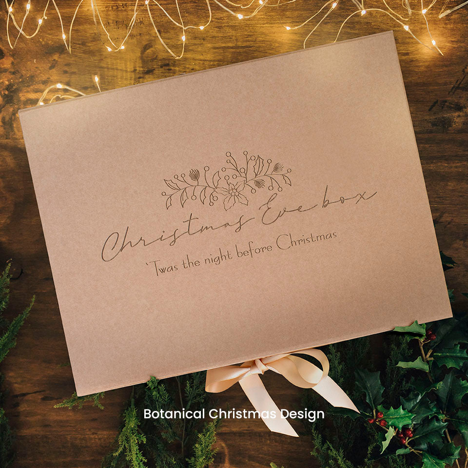 Hello Fern brown kraft gift box with 'Christmas Eve box' wording etched on lid and decorative holly on a wooden surface with greenery and lights.