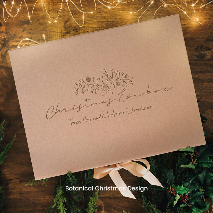Hello Fern brown kraft gift box with 'Christmas Eve box' wording etched on lid and decorative holly on a wooden surface with greenery and lights.