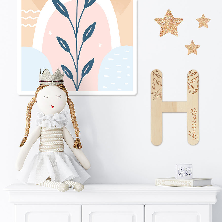 Closeup of styled wall in girl's bedroom with wooden Hello Fern customised wall letter 'H', doll, star stickers, and artwork all in neutral colours.