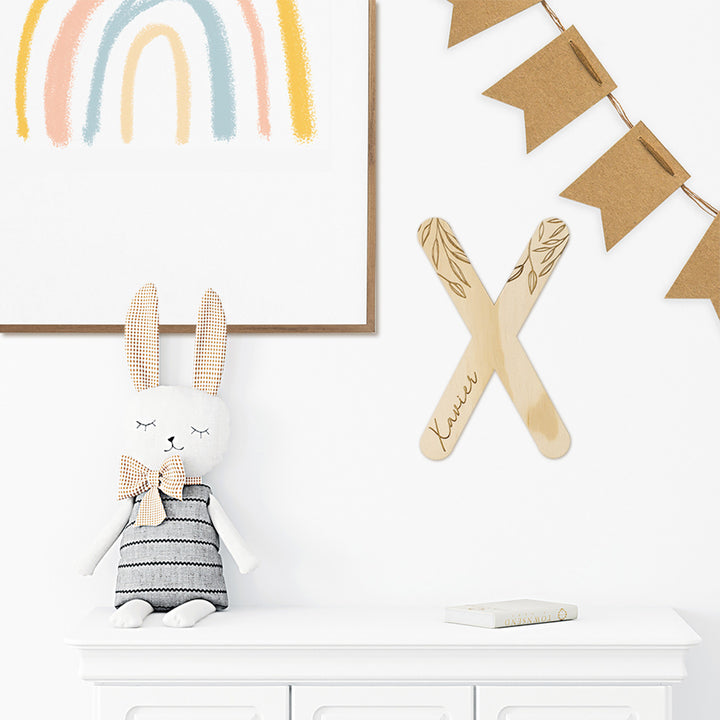 Closeup of styled wall in boy's bedroom with wooden Hello Fern customised wall letter 'X', stuffed animal rabbit, bunting, and child's rainbow artwork all in neutral colours.