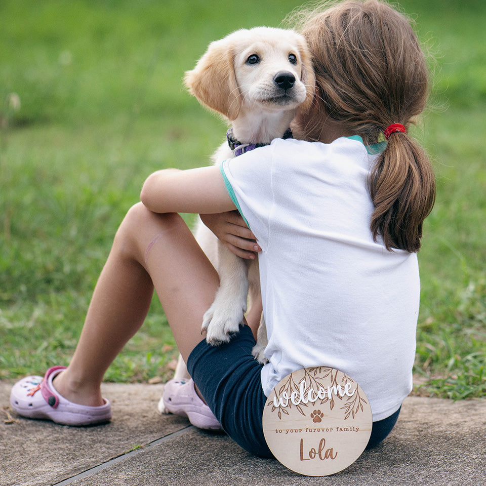 Little girl cuddling little puppy outside in grass with Hello Fern custom wooden pet welcome plaque.