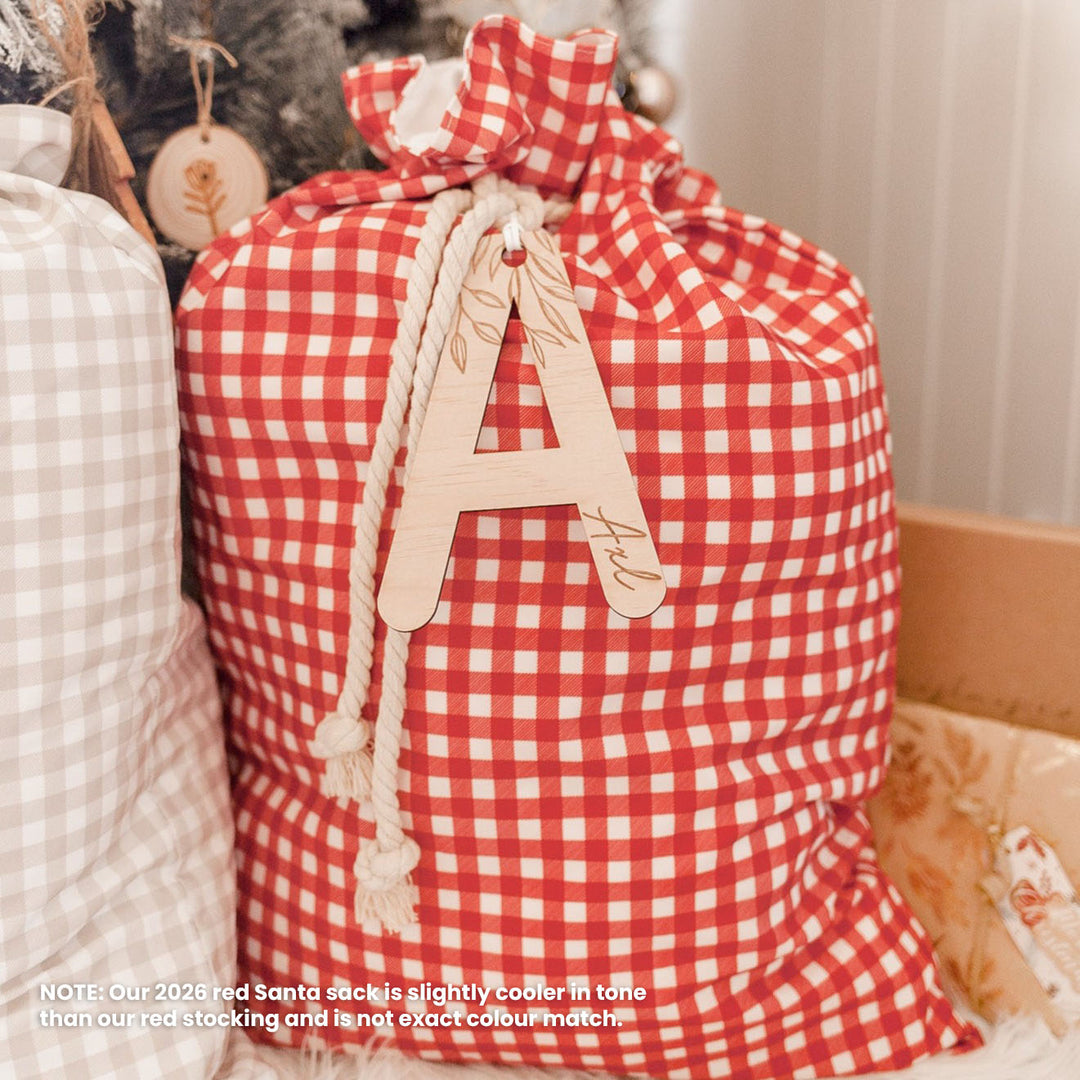 A red and white gingham check Christmas Santa sack with a drawstring closure, featuring a wooden letter 'A' monogram for 'Axl' hanging from the rope. A note in the bottom left corner explains that the red color is slightly cooler than expected.