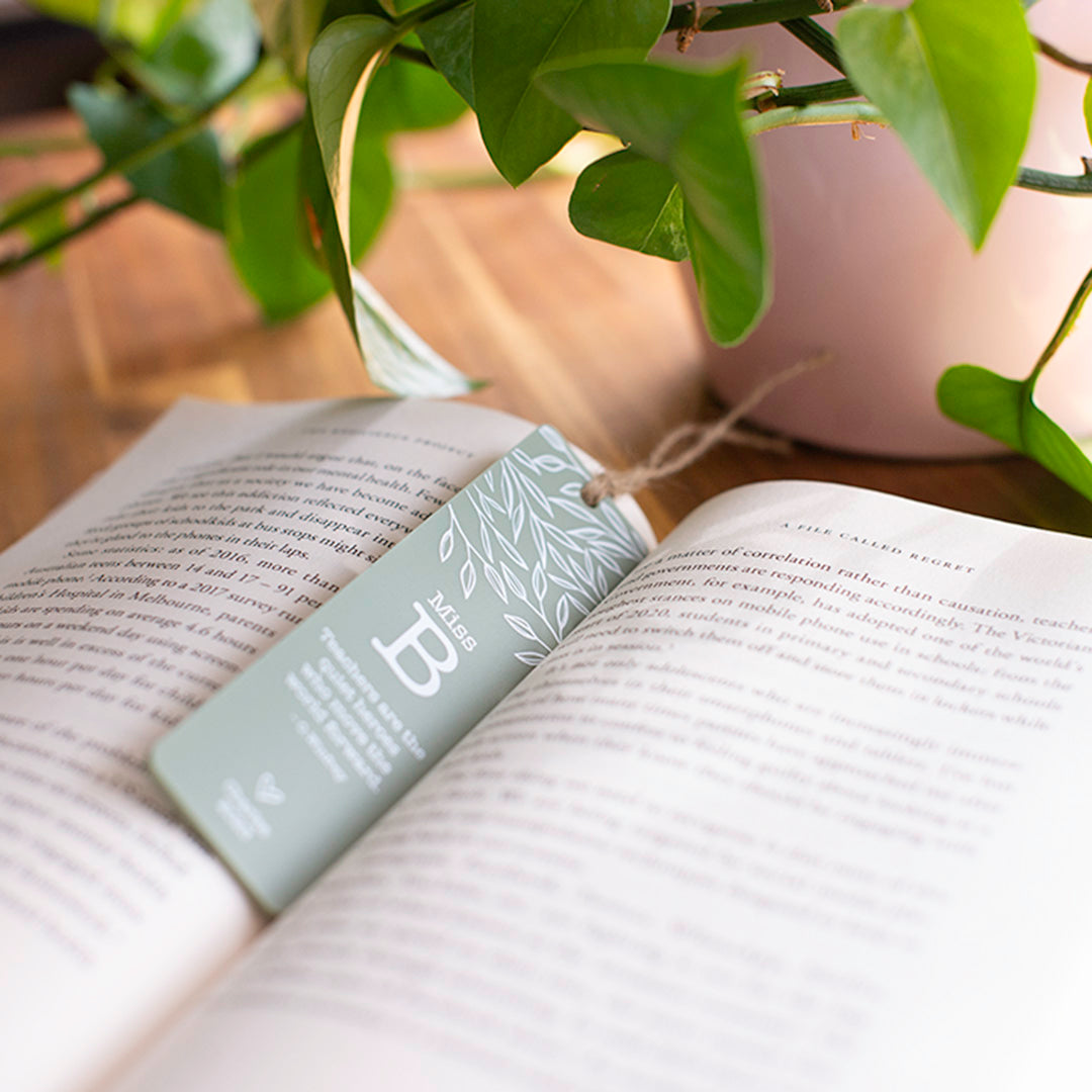 Photo of open novel with custom printed Hello Fern sage acrylic teacher bookmark in portrait orientation and plants in the background.