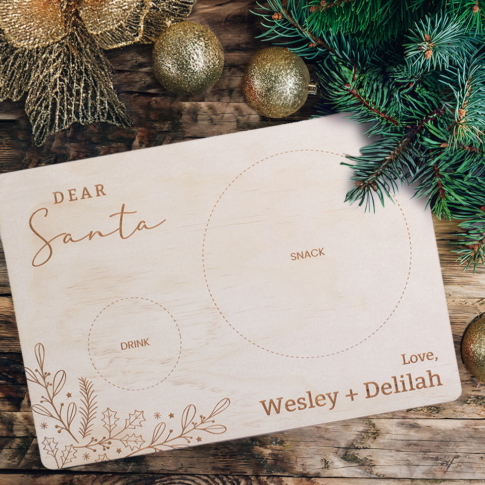 Overhead view of Hello Fern Santa Snack Board with custom names, snack and drink guides, and Christmas botanical design on wooden table with Christmas decor and pine branches.