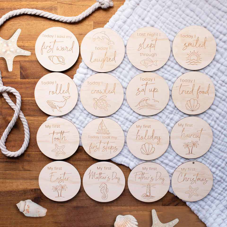 Set of 16 Hello Fern wooden baby achievement discs with engraved ocean-themed milestone phrases and graphics on a wooden surface with starfish and rope.
