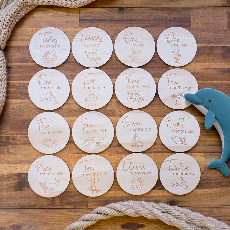 Set of 16 Hello Fern wooden baby month milestone discs with engraved ocean-themed graphics and wording on a wooden surface with a toy dolphin and rope.