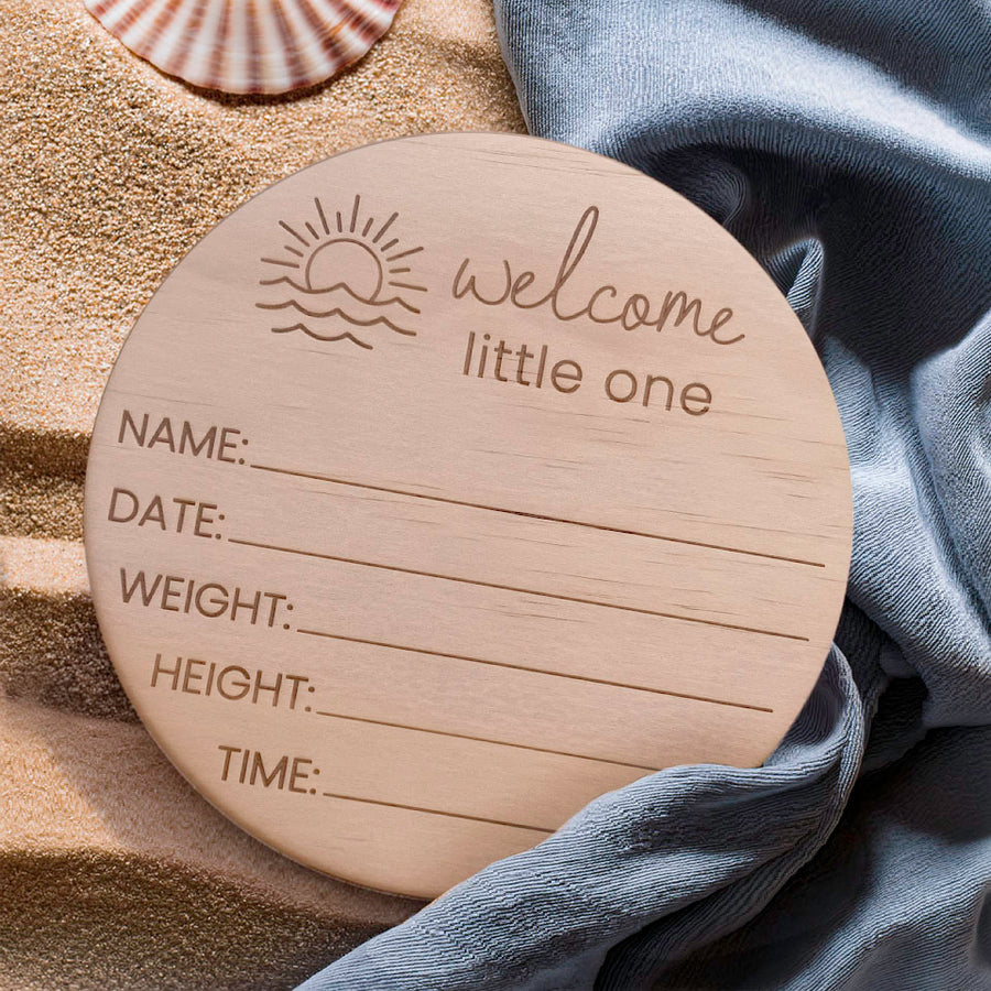Hello Fern round wooden baby birth announcement plaque with engraved text on a sandy background