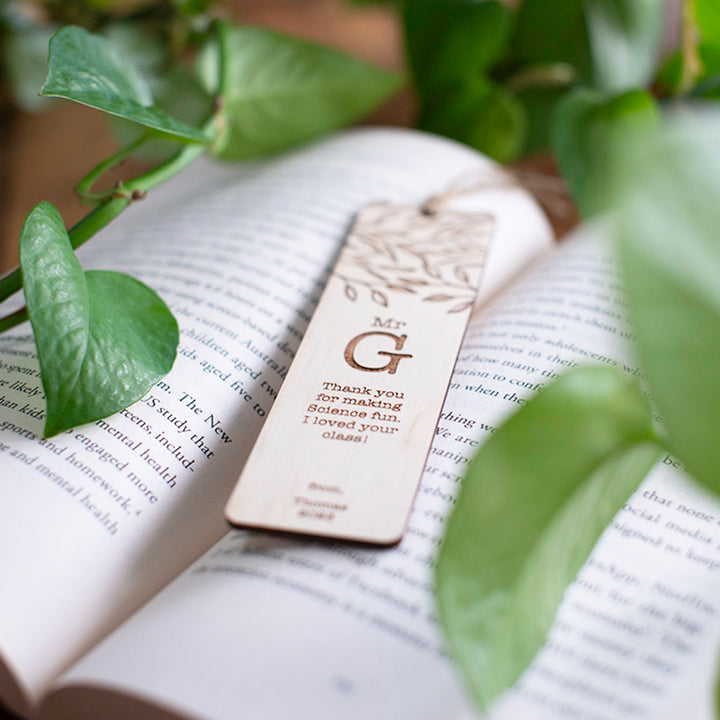 Photo of open book with custom etched Hello Fern portrait wooden teacher bookmark and plants in the background.