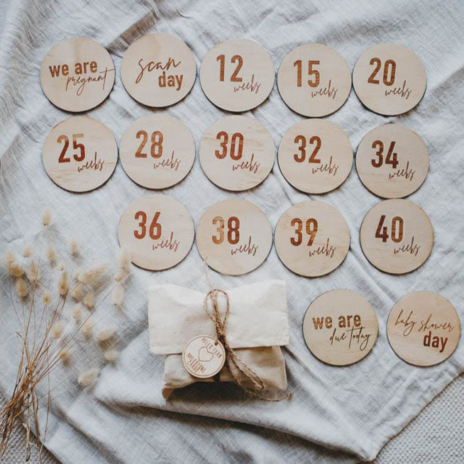 A collection of wooden pregnancy milestone cards with weeks marked on them, displayed on a neutral fabric surface, with a cotton bag and string on the side.