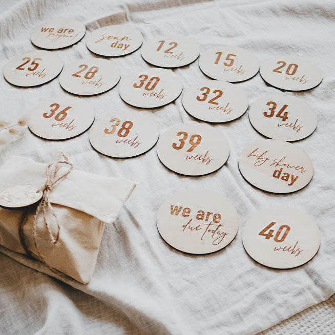 A collection of Hello Fern wooden pregnancy milestone wooden discs with "we are pregnant" to "40 weeks" and more displayed on a cloth background.