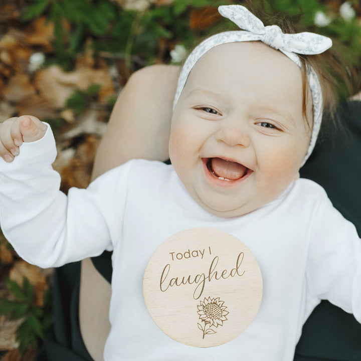 Happy little girl laughing on mother's lap with Hello Fern wooden country-themed achievement milestone disc "Today I laughed".