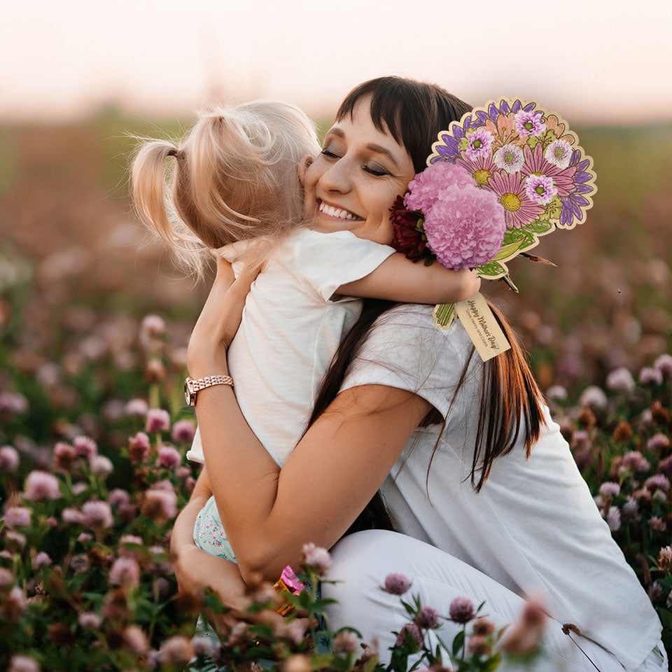 Mother and daughter embracing in hug in a field of wildflowers holding coloured-in Hello Fern Mother's Day custom wooden flower bouquet.