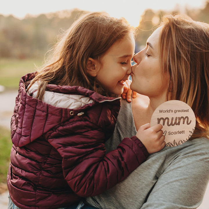 Mother holding and kissing daughter who is holding a Hello Fern custom wooden 'World's greatet mum' plaque for Mother's Day.