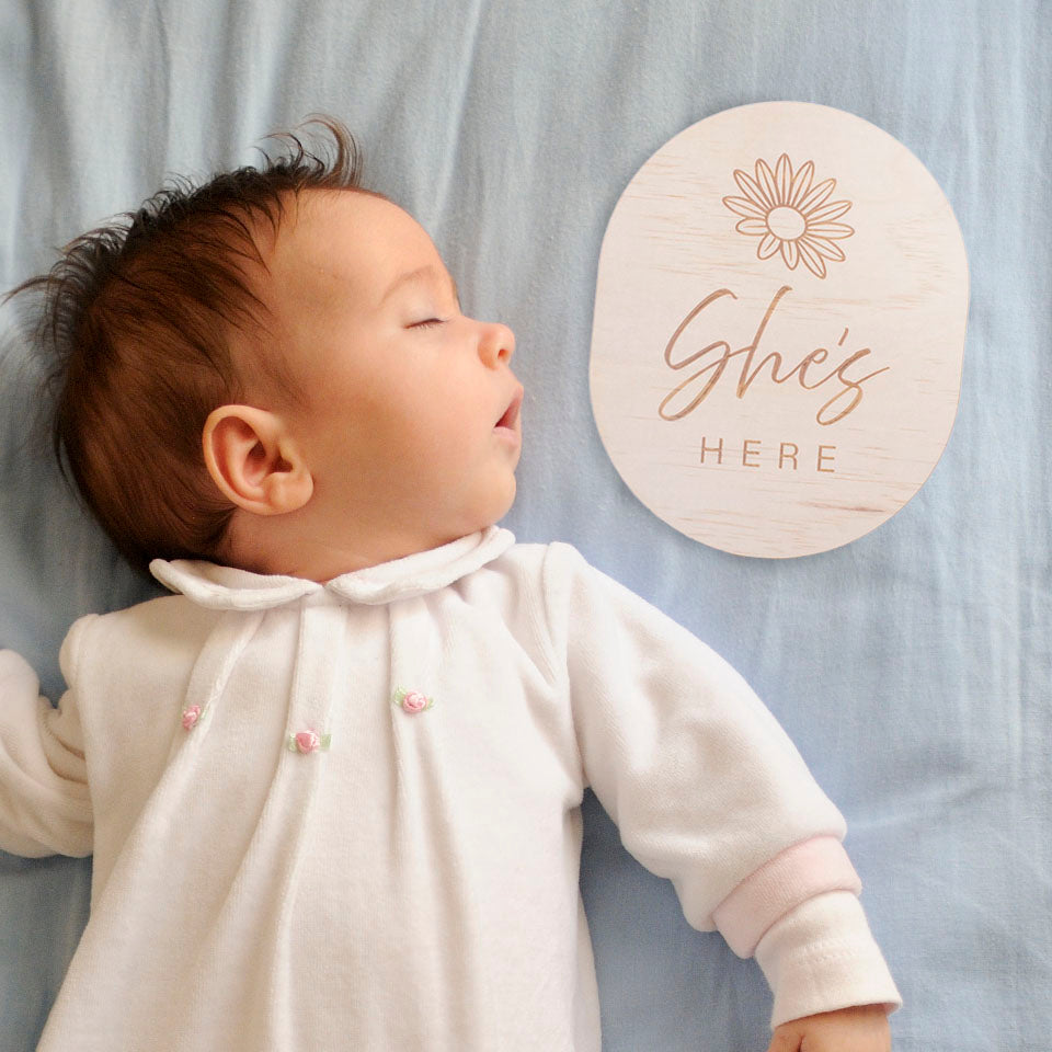 Sleeping newborn baby girl with Hello Fern double-sided wooden birth announcement plaque etched with a simple flower and the words "She's Here".