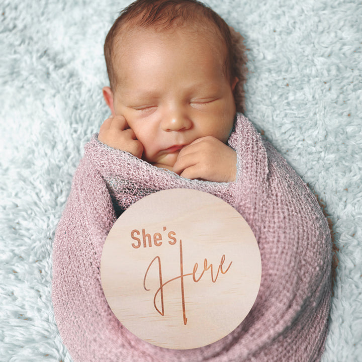 Newborn baby girl sleeping in swaddled blanket with Hello Fern timber double-sided birth announcement plaque in a classic design with the words "She's Here" etched.