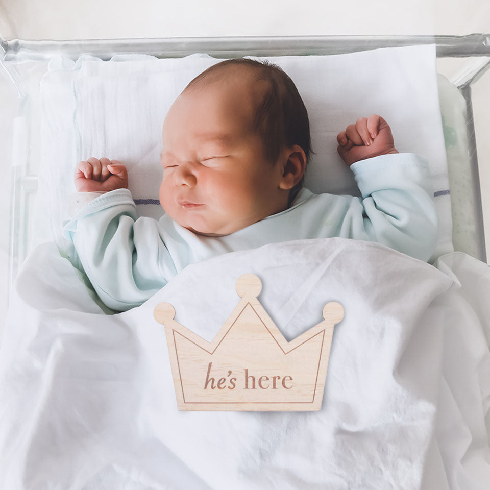 Newborn baby sleeping in hospital with Hello Fern timber "he's here" double-sided crown birth announcement plaque.