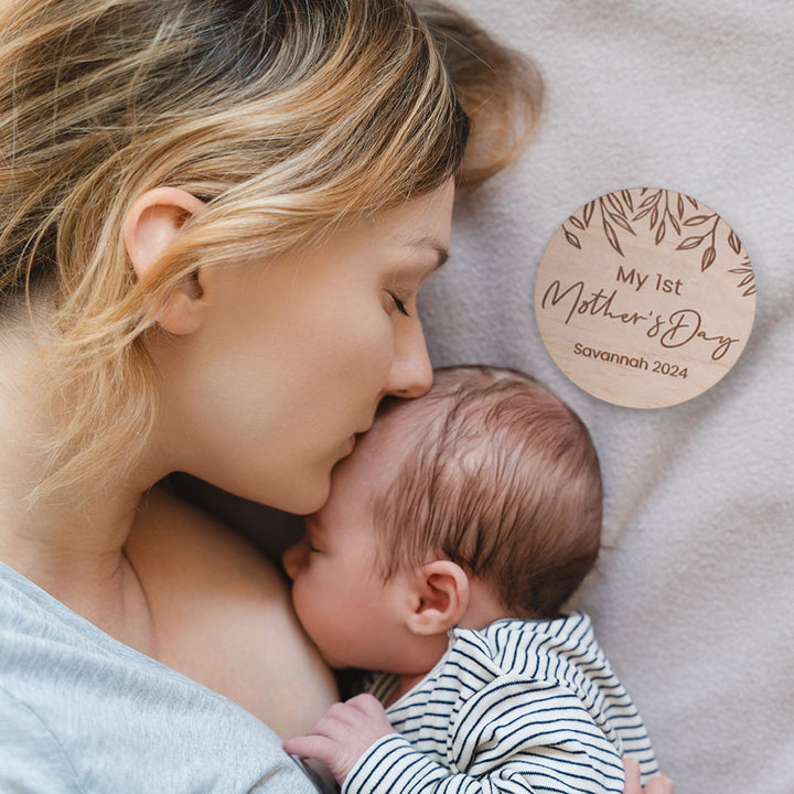 Overhead view of mother and baby snuggling in bed with "My 1st Mother's Day" wooden plaque.