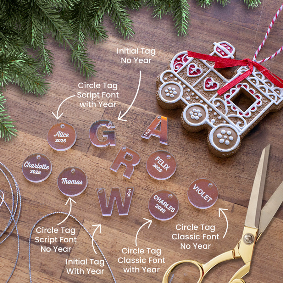 Christmas flat lay with various Hello Fern iridescent clear acrylic ornament tags including circle and inital tags personalised with child's name and various Christmas elements including train ornament, scissors, string, and pine branches.