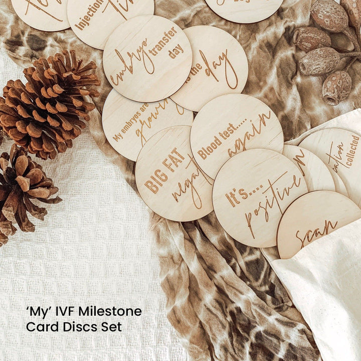 Hello Fern 'My' IVF milestone set of 15 wooden discs with inscriptions on a textured surface with pinecones.