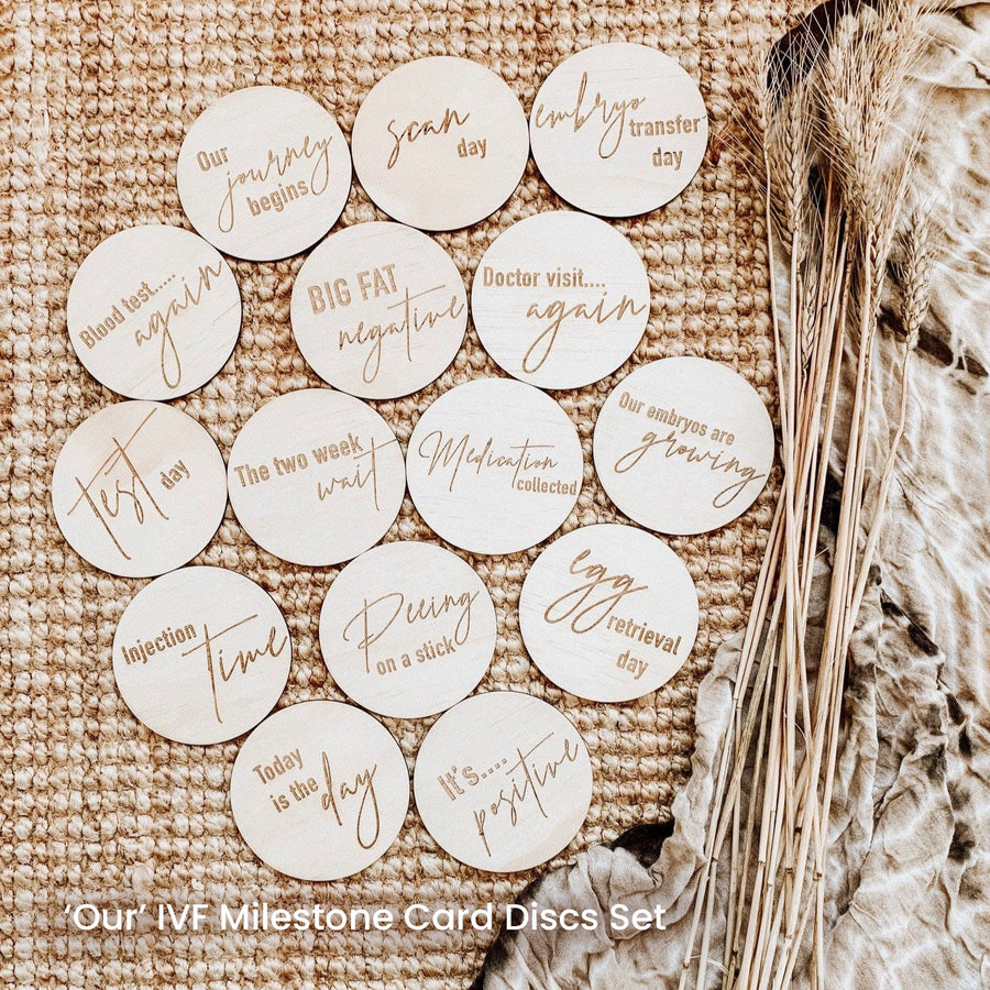 A collection of 15 Hello Fern wooden discs with 'Our' IVF milestone journey phrases written on them, laid out on a textured surface.