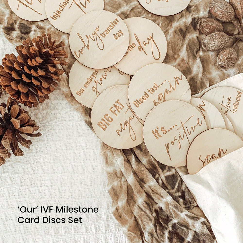 Hello Fern 'Our' IVF milestone set of 15 wooden discs with inscriptions on a textured surface with pinecones.