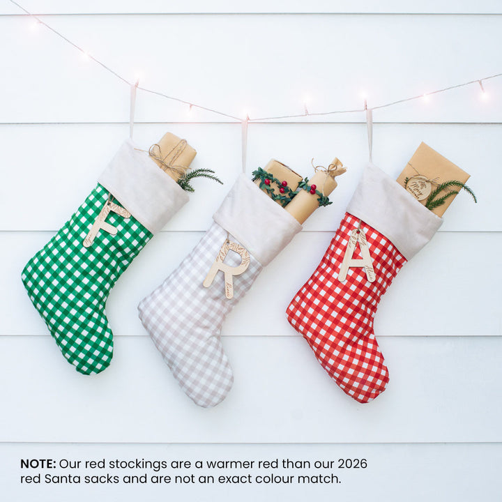 Three Christmas stockings hang on a white paneled wall under warm string lights. The left stocking is green and white gingham with a wooden letter F, the middle is white and beige gingham with a wooden letter R, and the right is red and white gingham with a wooden letter A.