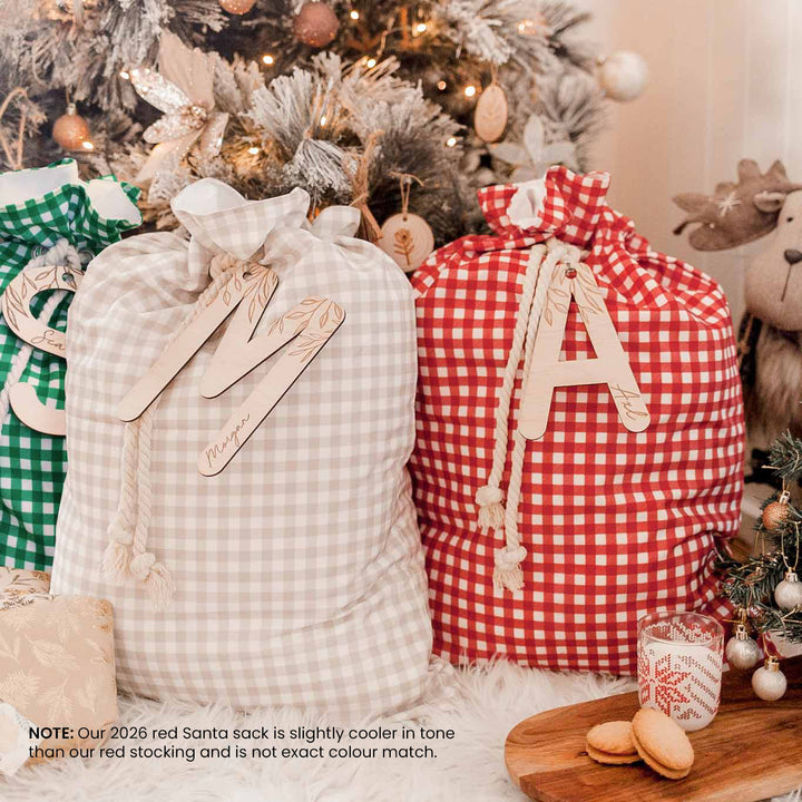Three personalised Christmas Santa sacks with gingham fabric and wooden monogram letters 'M', 'A', and 'S', styled in front of a decorated Christmas tree with a note explaining the red bag's slightly cooler tone.