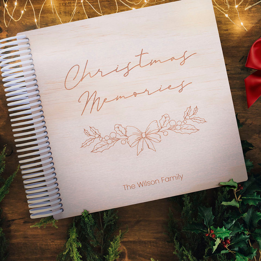 Top view of Hello Fern's wooden custom Christmas photo album. Cover custom-etched with Christmas holly and bow and wording 'Christmas Memories - The Wilson Family' which can be personalised. Christmas fairy lights, holly, and red bow surrounding on a wooden table top.