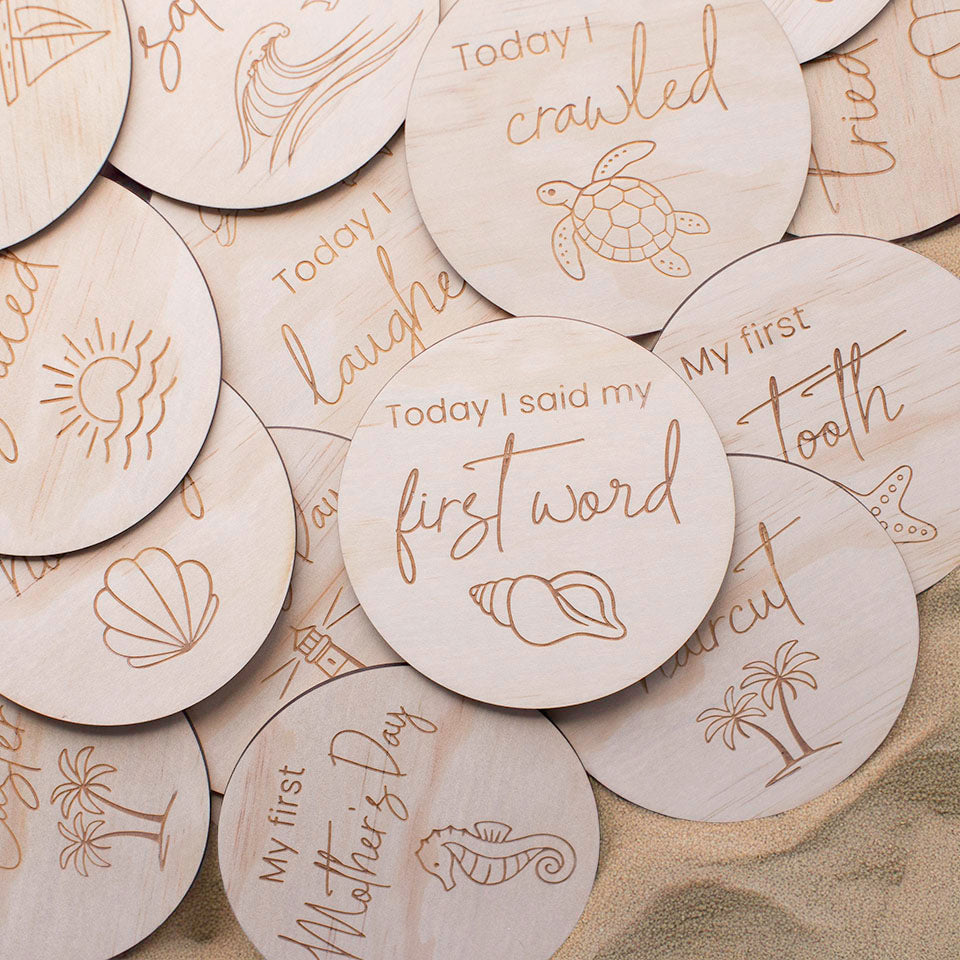 Close up of Hello Fern wooden baby achievement ocean-themed milestone discs with engraved text and illustrations on a sand background.