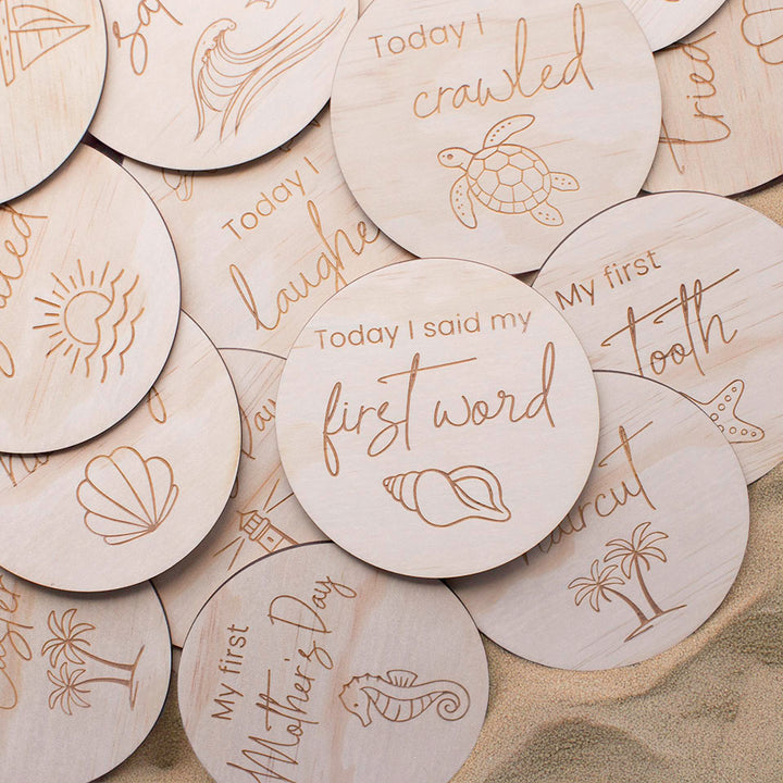Close up of Hello Fern wooden baby achievement ocean-themed milestone discs with engraved text and illustrations on a sand background.