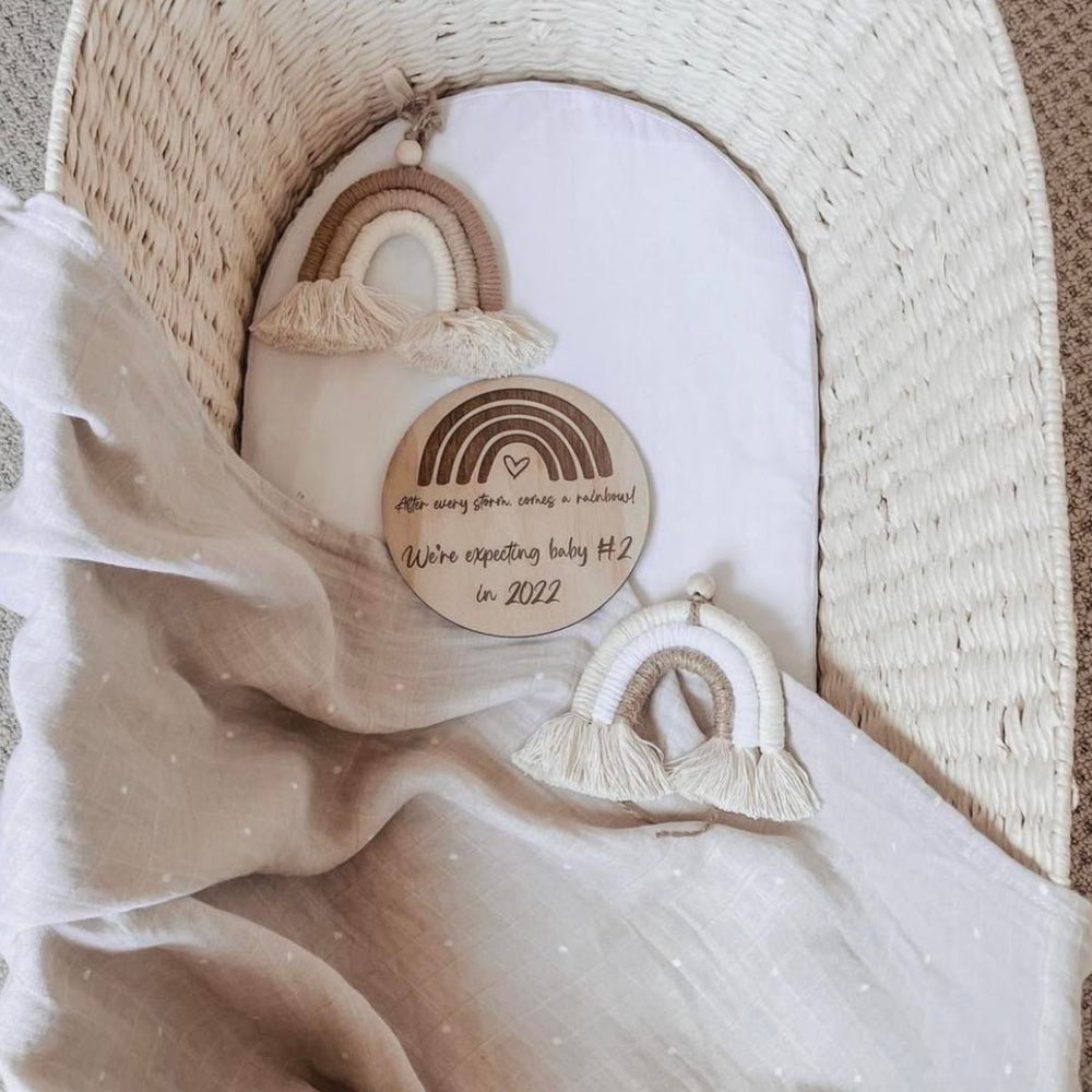 Hello Fern wooden announcement disc in rainbow design with custom text shown in a baby bassinet.