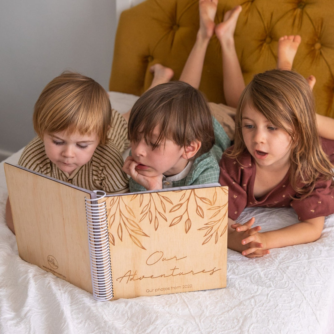 Three children lying on bed looking through Hello Fern wooden family photo album.
