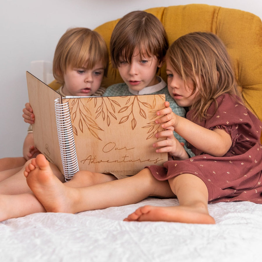 Three children sitting on bed looking through a Hello Fern wooden photo album of family adventures.