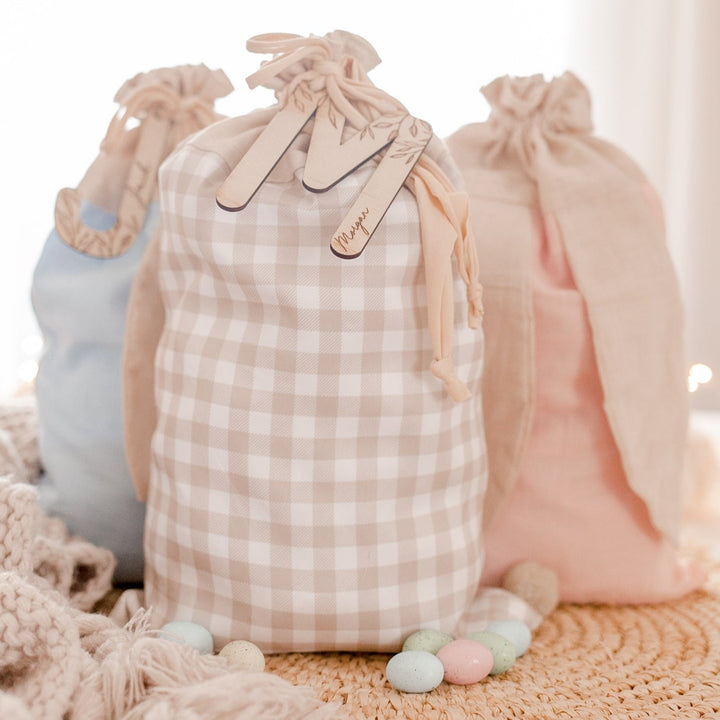 Three cotton Easter sacks (beige gingham, blue and pink) with custom wooden letter name plaque sitting on table with Easter eggs.